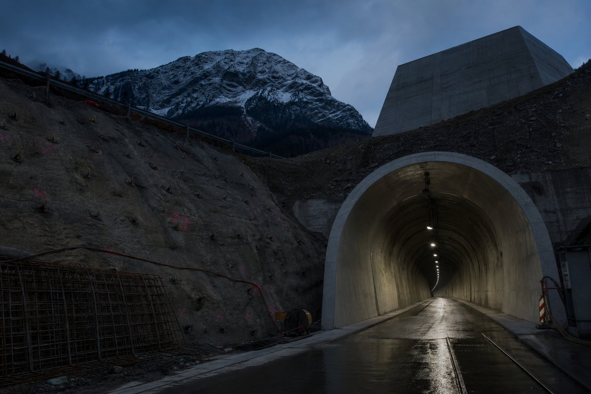 Archival-feeling engineering scene, dim and documentary: a mountainous transport corridor, at blue hour with the portal lit, shot at 35mm. (AI-generated image)