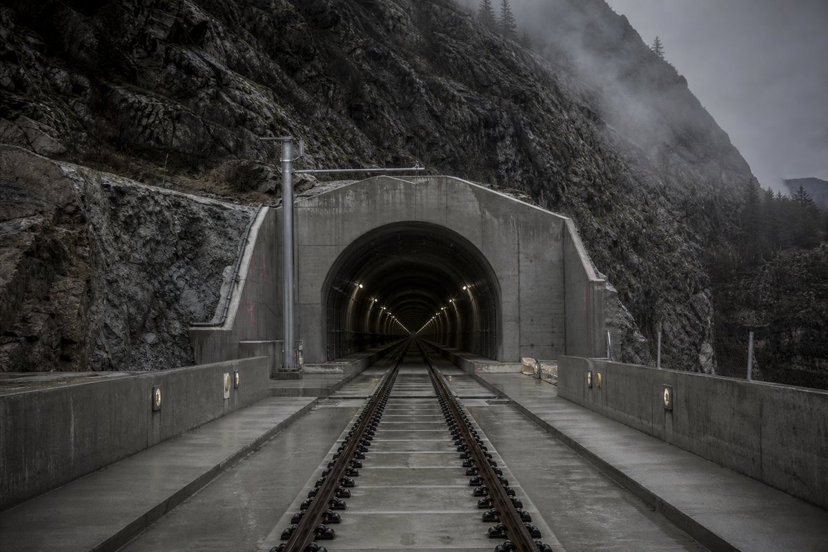 Archival-feeling engineering scene, dim and documentary: a mountainous transport corridor, in drifting mist, a dead-centre symmetrical composition. (AI-generated image)