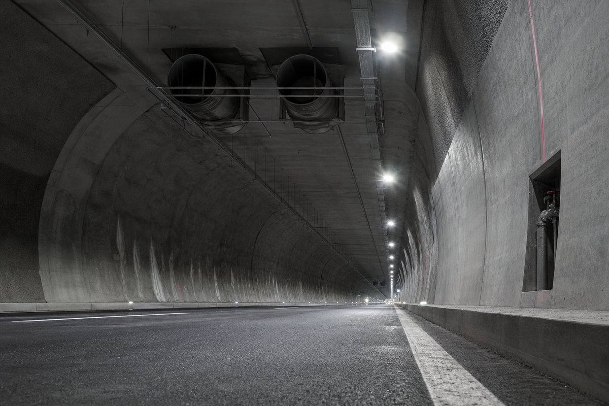 Interior of a long two-lane Austrian motorway tunnel with bidirectional traffic: a single wide bore, transverse ventilation ducts above the false ceiling. (AI-generated image)