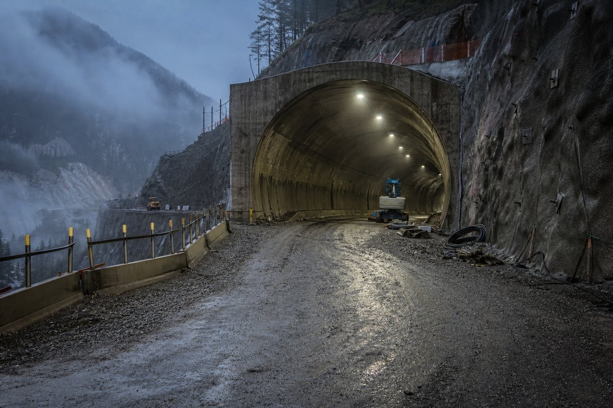 Archival-feeling engineering scene, dim and documentary: a mountainous transport corridor, at dusk with lights just on, shot at 50mm, compressed. (AI-generated image)