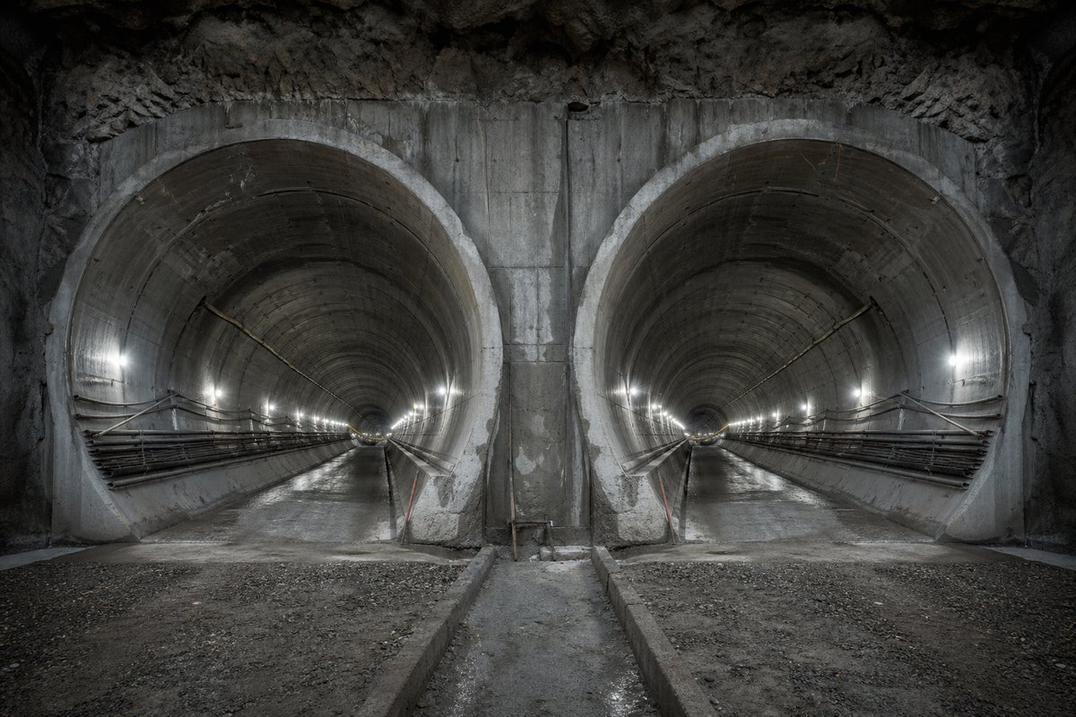Two parallel tunnel bores diverging behind a shared portal wall, cold LED lighting, in flat overcast light, a dead-centre symmetrical composition. Frame 490. (AI-generated image)