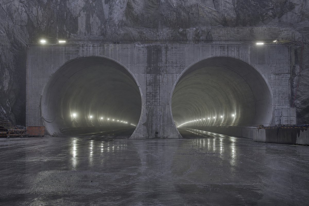 Two parallel tunnel bores diverging behind a shared portal wall, cold LED lighting, in thin fog, shot at 85mm from a distance. Frame 700. (AI-generated image)