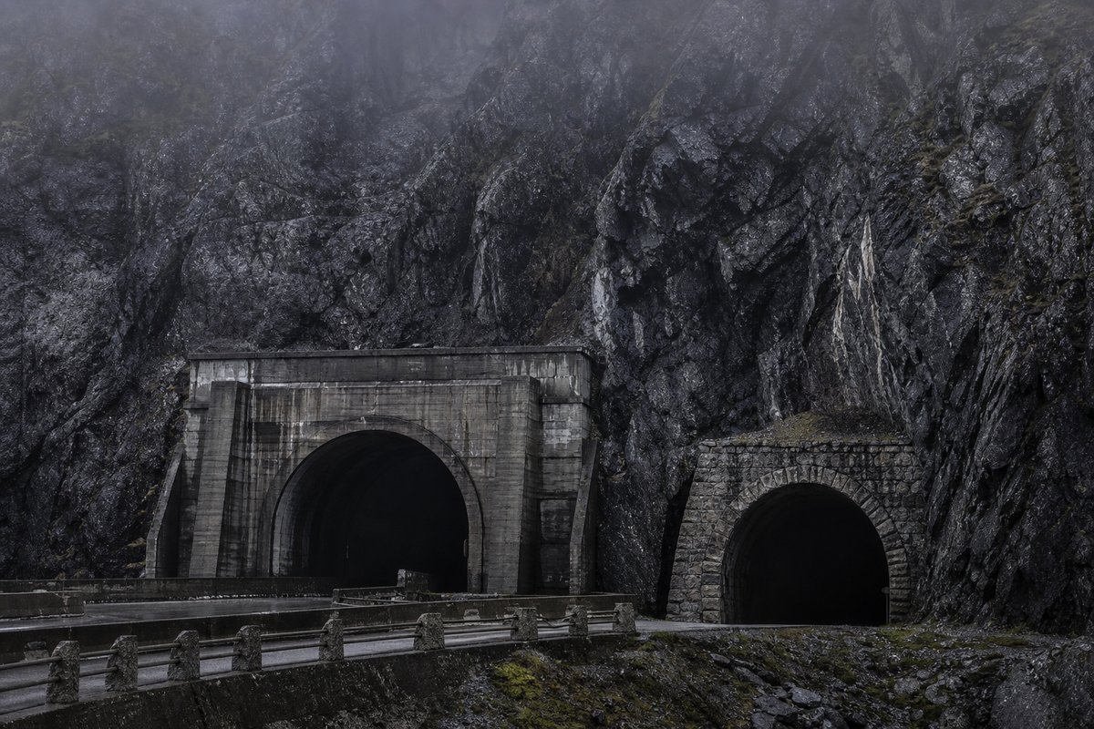 Rock face carrying two tunnel mouths of visibly different generations, wet stone, in thin fog, shot at 85mm from a distance. Frame 689. (AI-generated image)