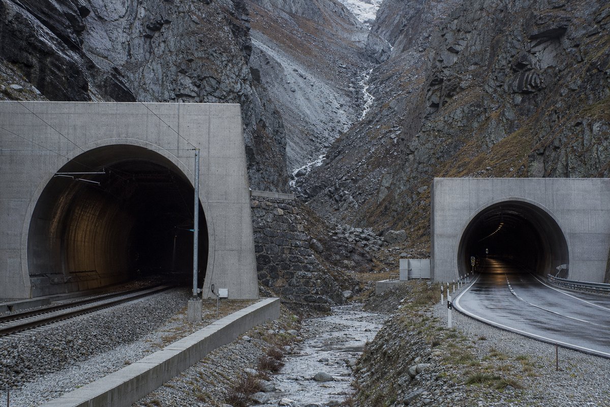 Railway portal and a road portal facing each other across a narrow valley floor, in cold clear light, shot at 50mm. Frame 858. (AI-generated image)