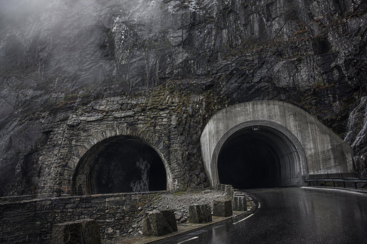 Rock face carrying two tunnel mouths of visibly different generations, wet stone, in drifting mist, shot at 35mm. Frame 965. (AI-generated image)
