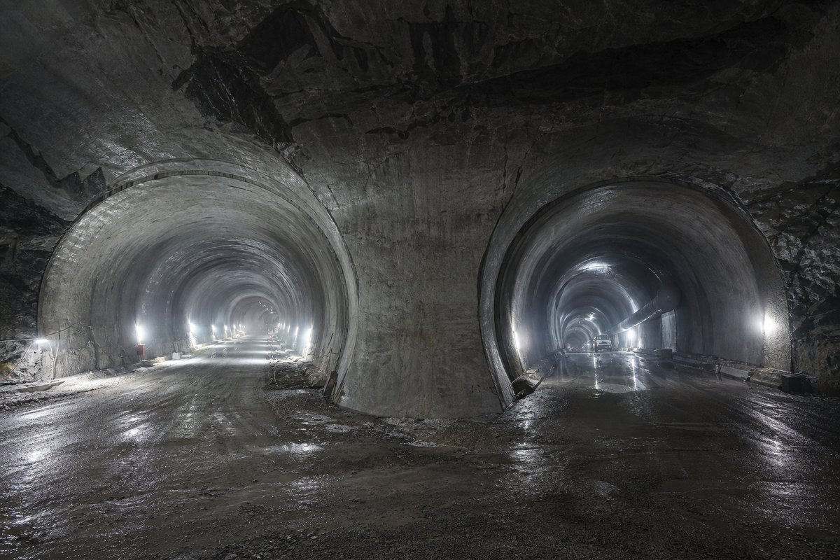 Two parallel tunnel bores diverging behind a shared portal wall, cold LED lighting, in drifting mist, shot wide at 24mm. Frame 486. (AI-generated image)