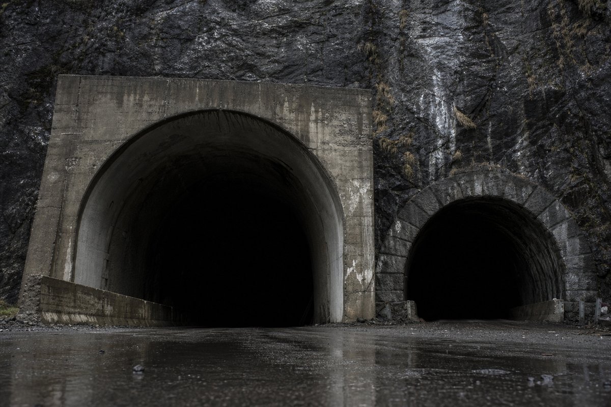 Rock face carrying two tunnel mouths of visibly different generations, wet stone, in flat overcast light, a low camera position. Frame 609. (AI-generated image)