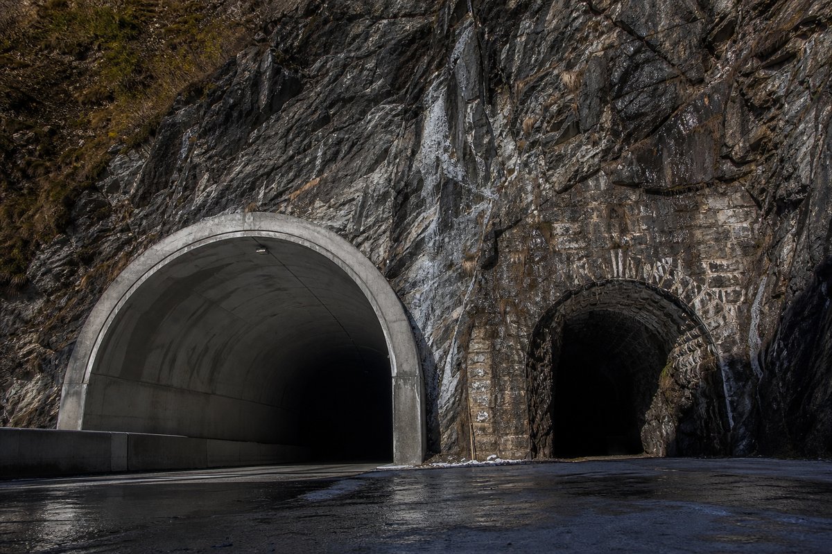 Rock face carrying two tunnel mouths of visibly different generations, wet stone, in hard low winter sun, a low camera position. Frame 104. (AI-generated image)