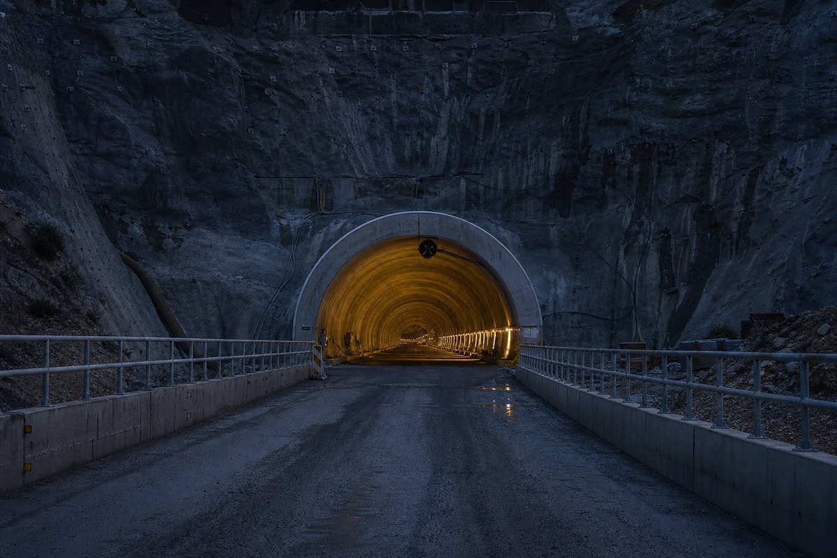 Mouth of a long bore seen from the approach cutting, guardrails converging, at blue hour with the portal lit, shot at 85mm from a distance. Frame 51. (AI-generated image)
