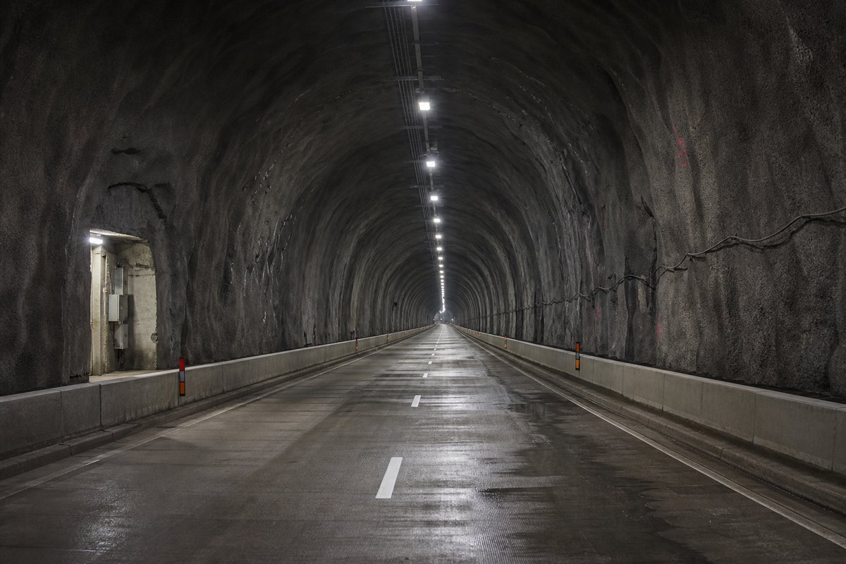Looking down the axis of an extremely long two-lane road tunnel bore, luminaires receding to a point: a mountainous transport corridor, in hard low winter sun, shot at 50mm. (AI-generated image)