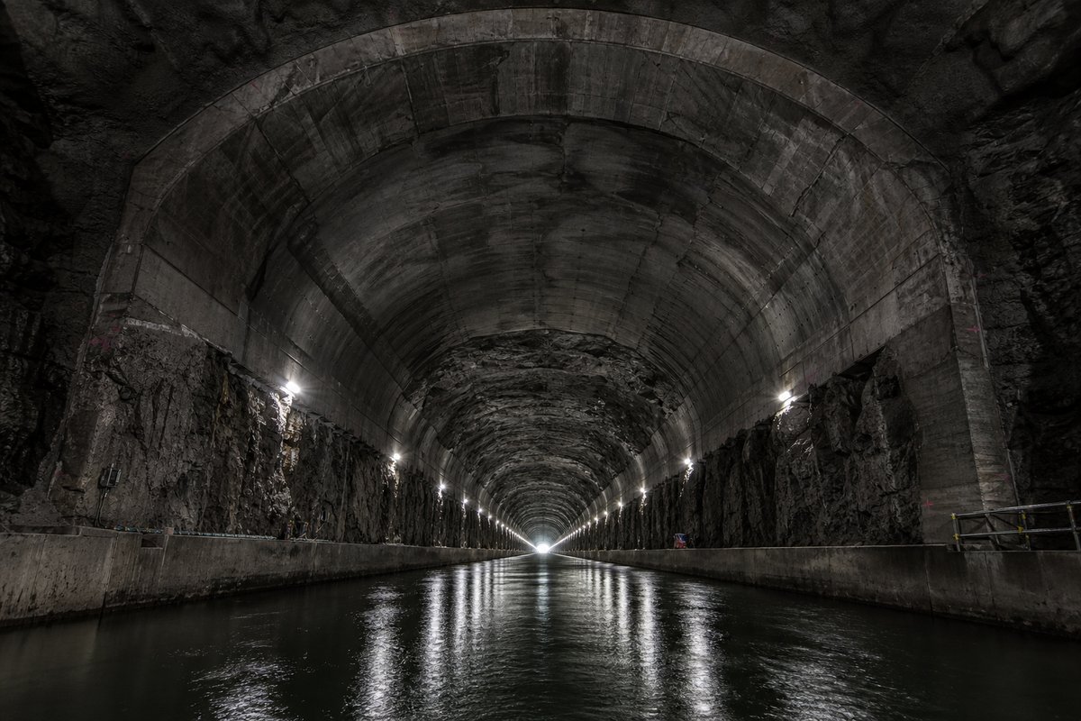 Narrow stone-lined canal tunnel over dark still water, the far end a small bright arch: a mountainous transport corridor, under a heavy dark sky. (AI-generated image)