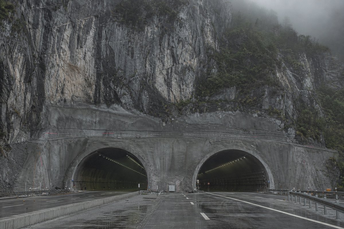 Road tunnel portal in China: karst limestone towers in mist, a broad new expressway portal cut into the cliff, in cold clear light, shot at 50mm, compressed. (AI-generated image)