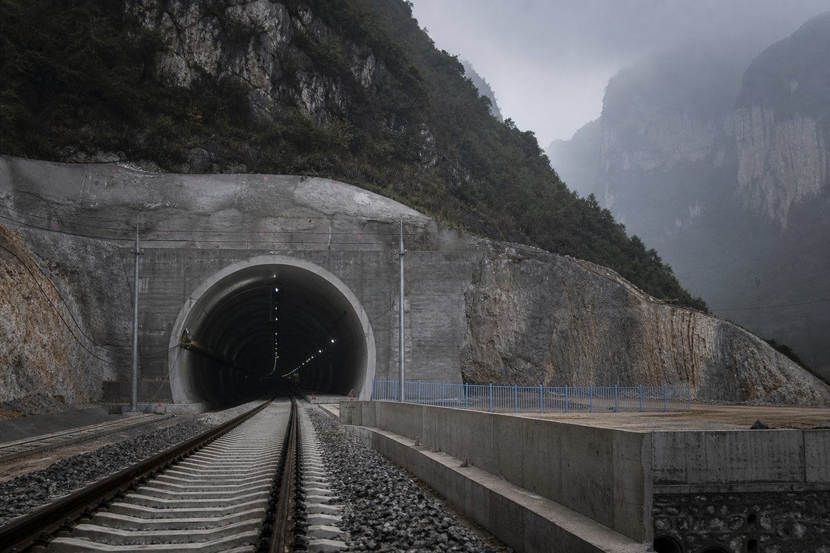 Railway tunnel portal with track running in in China: karst limestone towers in mist, a broad new expressway portal cut into the cliff, in thin morning fog, shot at 50mm. (AI-generated image)