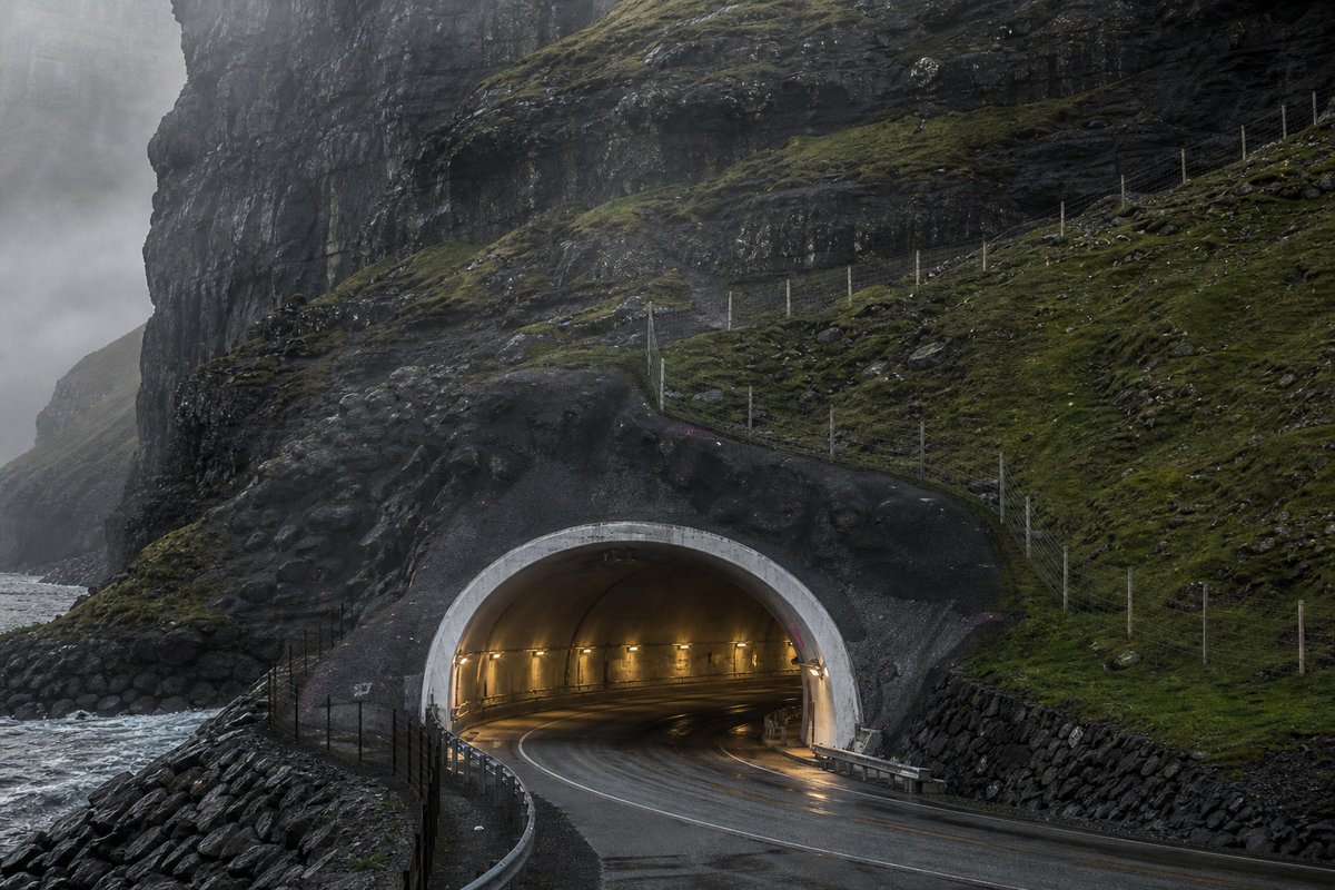 Road tunnel portal in Faroe Islands: sheer sea cliff and grass, the mouth of a subsea tunnel, in drifting mist, shot at 85mm from a distance. (AI-generated image)