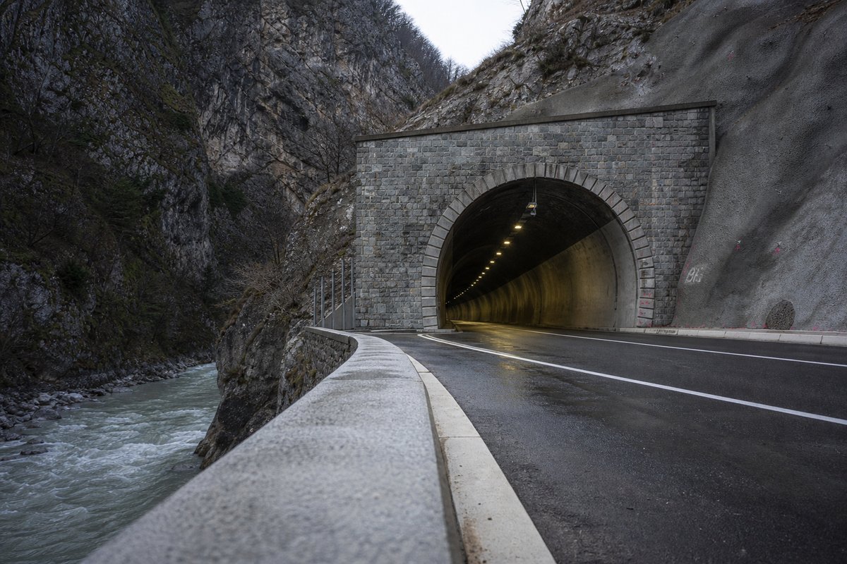 Transport infrastructure structure in France: Alpine gorge with a stone-faced tunnel mouth beside a river, in hard low winter sun, a low camera position near the road surface. (AI-generated image)