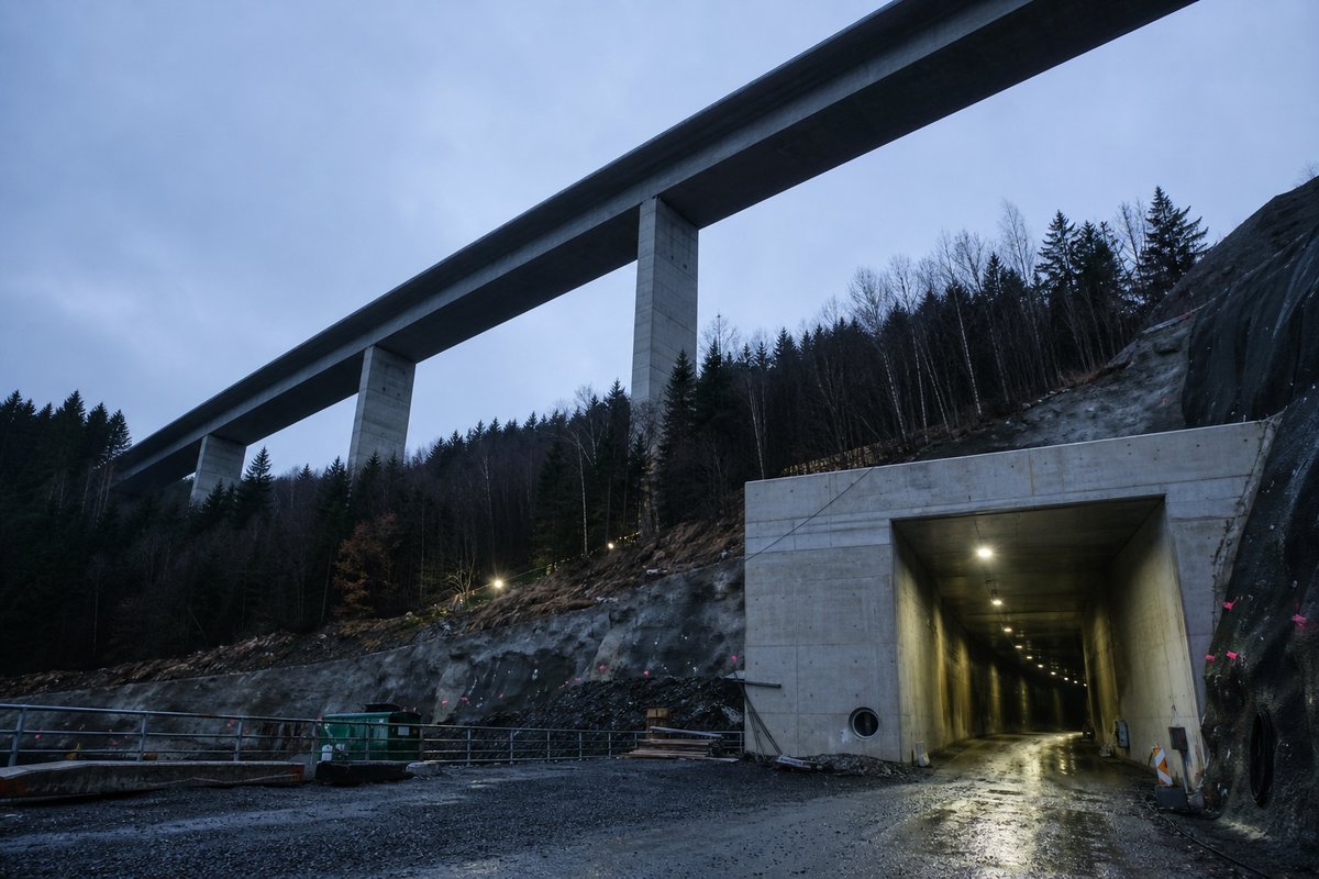 Long concrete box-girder viaduct on tall piers, seen from below in Germany: wooded low mountain range, a cut-and-cover portal in fair-faced concrete, at dusk with lights just on. (AI-generated image)