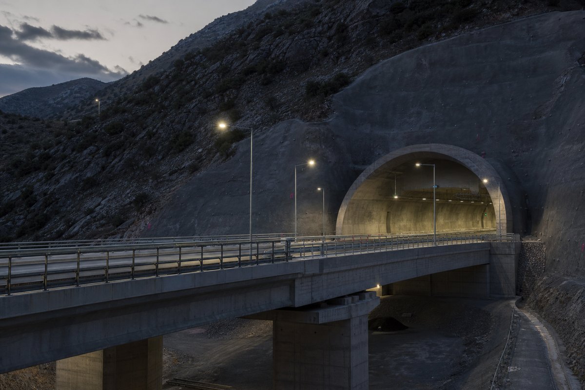 Road tunnel portal in Greece: dry mountain scrub, a motorway portal above a viaduct deck, at dusk with lights just on, shot wide at 24mm. (AI-generated image)