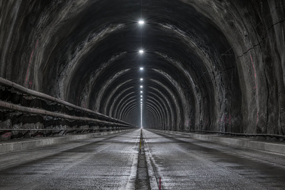Looking down the axis of an extremely long two-lane road tunnel bore, luminaires receding to a point: a mountainous transport corridor, in cold clear light. (AI-generated image)