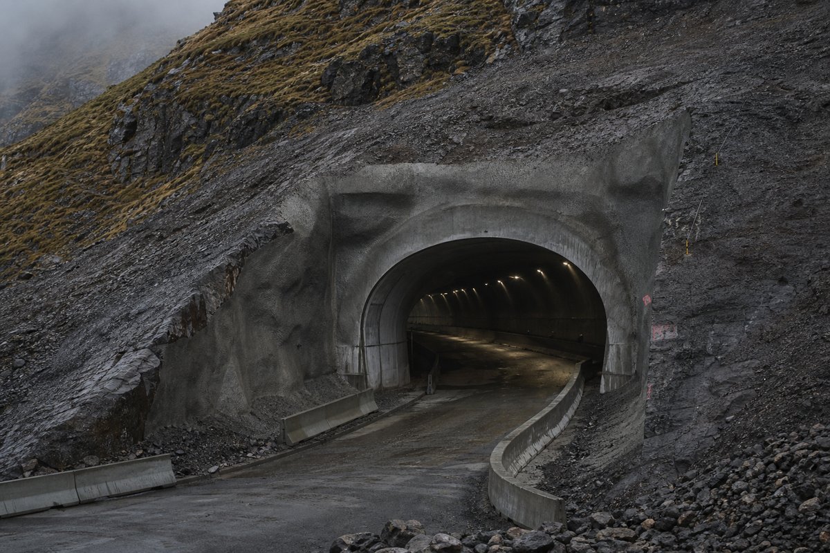 Road tunnel portal in Iceland: black basalt and moss, a raw rock tunnel mouth under low cloud, in flat overcast light, a slightly elevated three-quarter view. (AI-generated image)