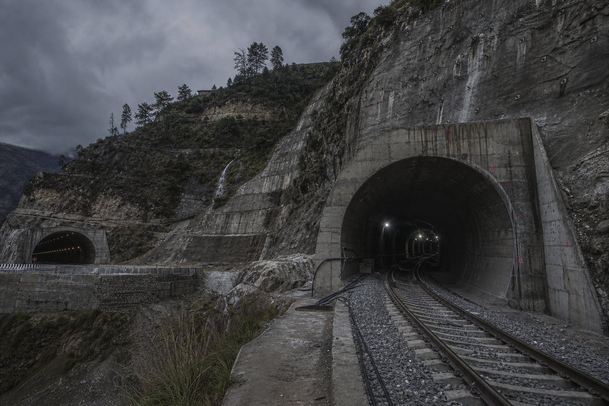 Railway tunnel portal with track running in in India: Himalayan slope with terraced switchbacks and a new highway portal, under a heavy dark sky, shot at 35mm. (AI-generated image)