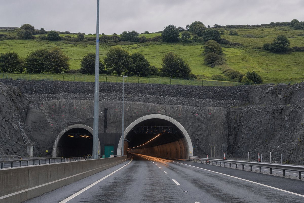 Road tunnel portal in Ireland: green drumlin landscape with a motorway tunnel portal, in flat overcast light, shot at 50mm, compressed. (AI-generated image)