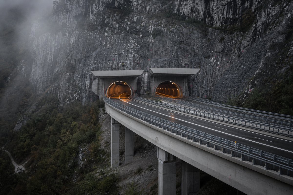 Motorway viaduct and tunnel portal in sequence along an autostrada in Italy: Apennine limestone ridge, twin concrete motorway portals with a projecting canopy, in thin morning fog. (AI-generated image)