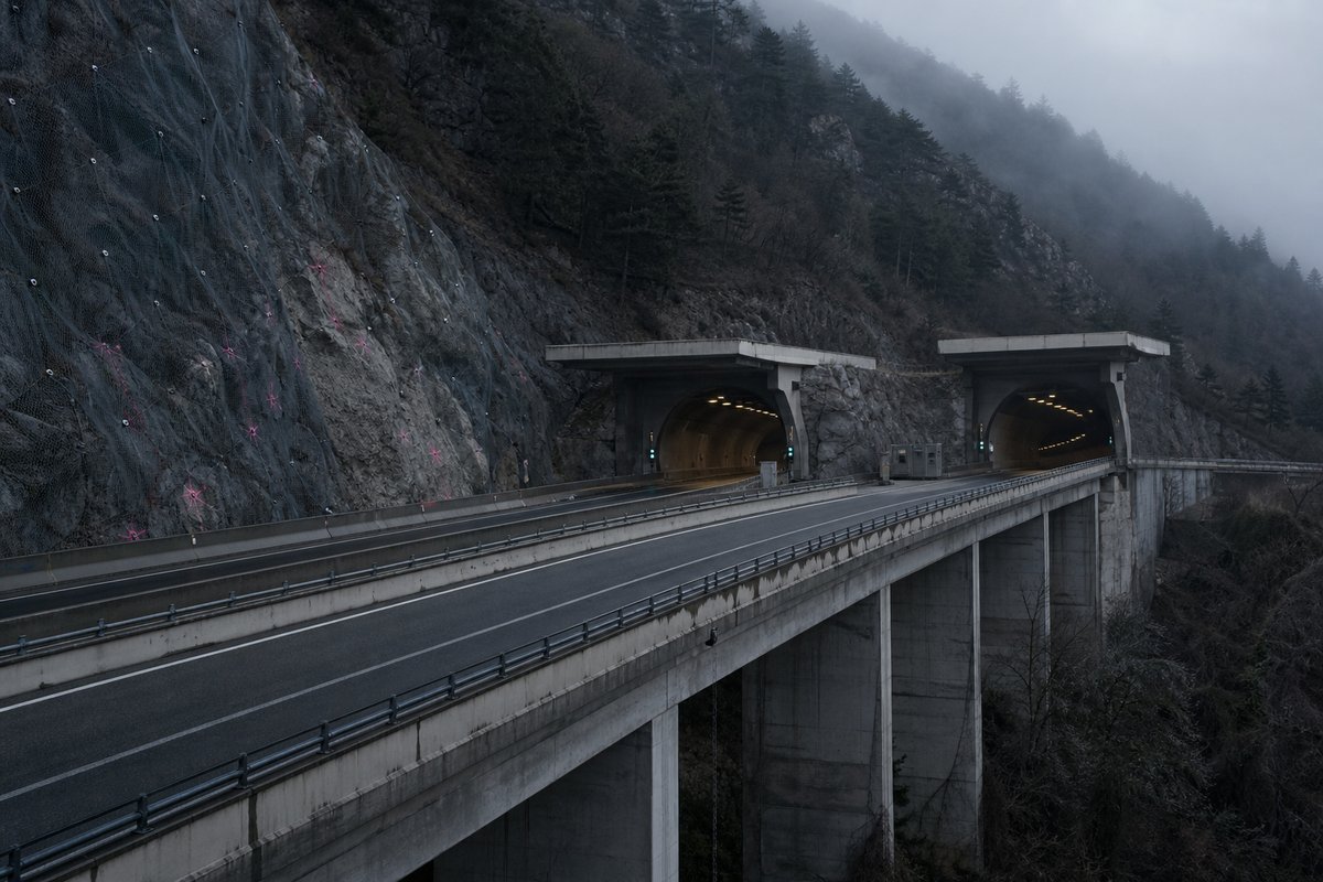 Motorway viaduct and tunnel portal in sequence along an autostrada in Italy: Apennine limestone ridge, twin concrete motorway portals with a projecting canopy, in thin morning fog. (AI-generated image)
