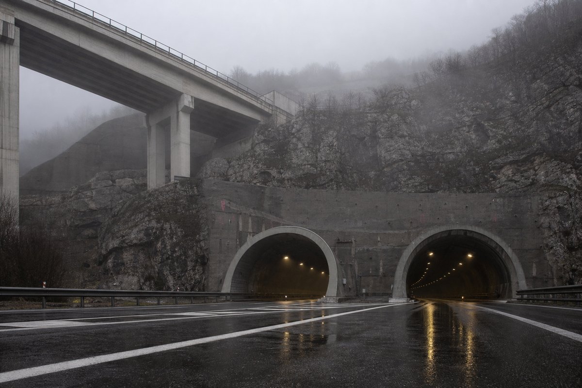 Motorway viaduct and tunnel portal in sequence along an autostrada in Italy: Apennine limestone ridge, twin concrete motorway portals with a projecting canopy, in thin morning fog. (AI-generated image)