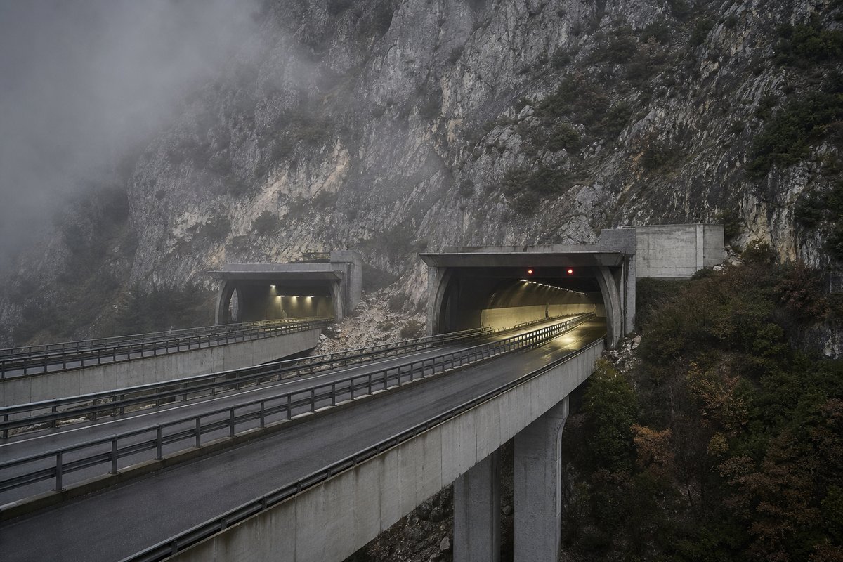 Motorway viaduct and tunnel portal in sequence along an autostrada in Italy: Apennine limestone ridge, twin concrete motorway portals with a projecting canopy, in drifting mist. (AI-generated image)