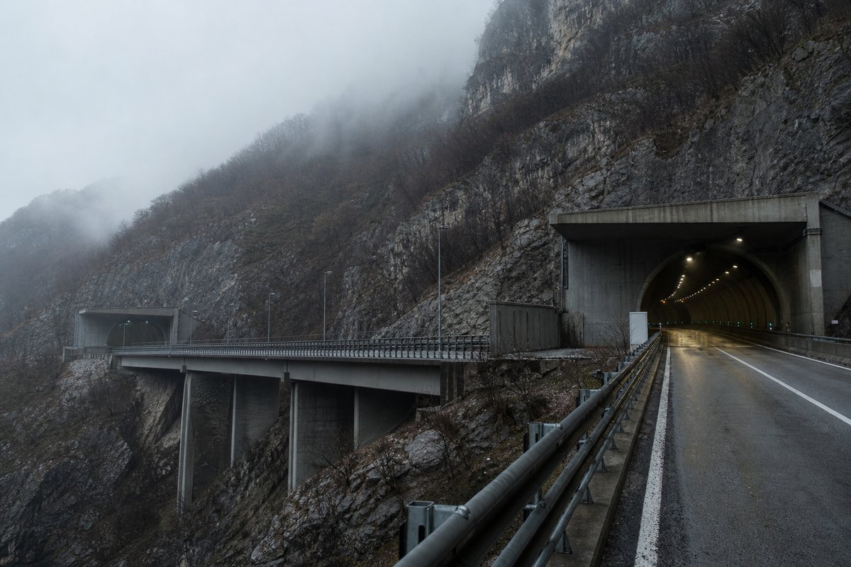 Motorway viaduct and tunnel portal in sequence along an autostrada in Italy: Apennine limestone ridge, twin concrete motorway portals with a projecting canopy, in thin morning fog. (AI-generated image)