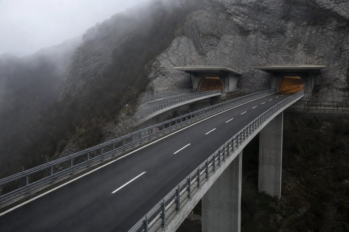 Motorway viaduct and tunnel portal in sequence along an autostrada in Italy: Apennine limestone ridge, twin concrete motorway portals with a projecting canopy, in drifting mist. (AI-generated image)