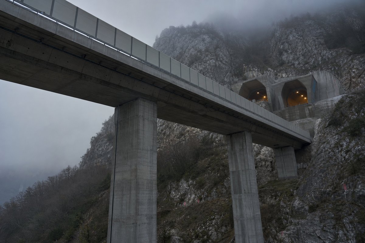 Long concrete box-girder viaduct on tall piers, seen from below in Italy: Apennine limestone ridge, twin concrete motorway portals with a projecting canopy, in thin morning fog. (AI-generated image)