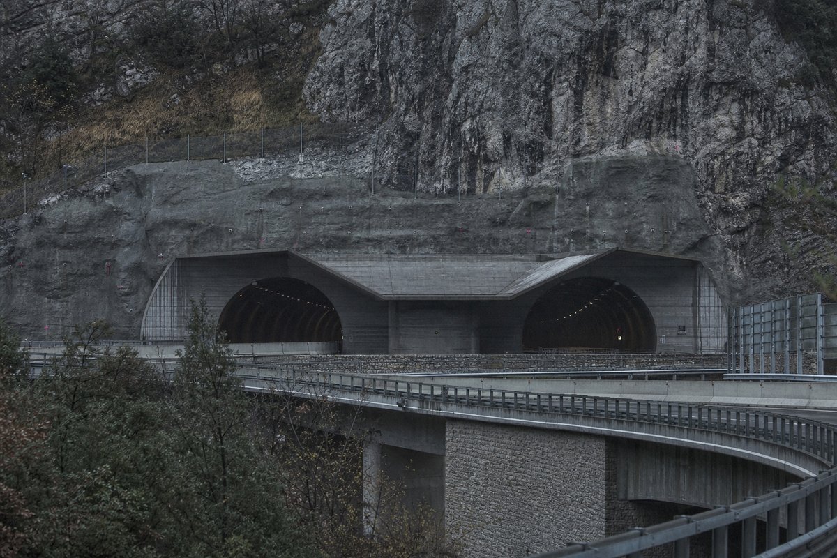 Transport infrastructure structure in Italy: Apennine limestone ridge, twin concrete motorway portals with a projecting canopy, in flat overcast light, shot at 85mm from a distance. (AI-generated image)