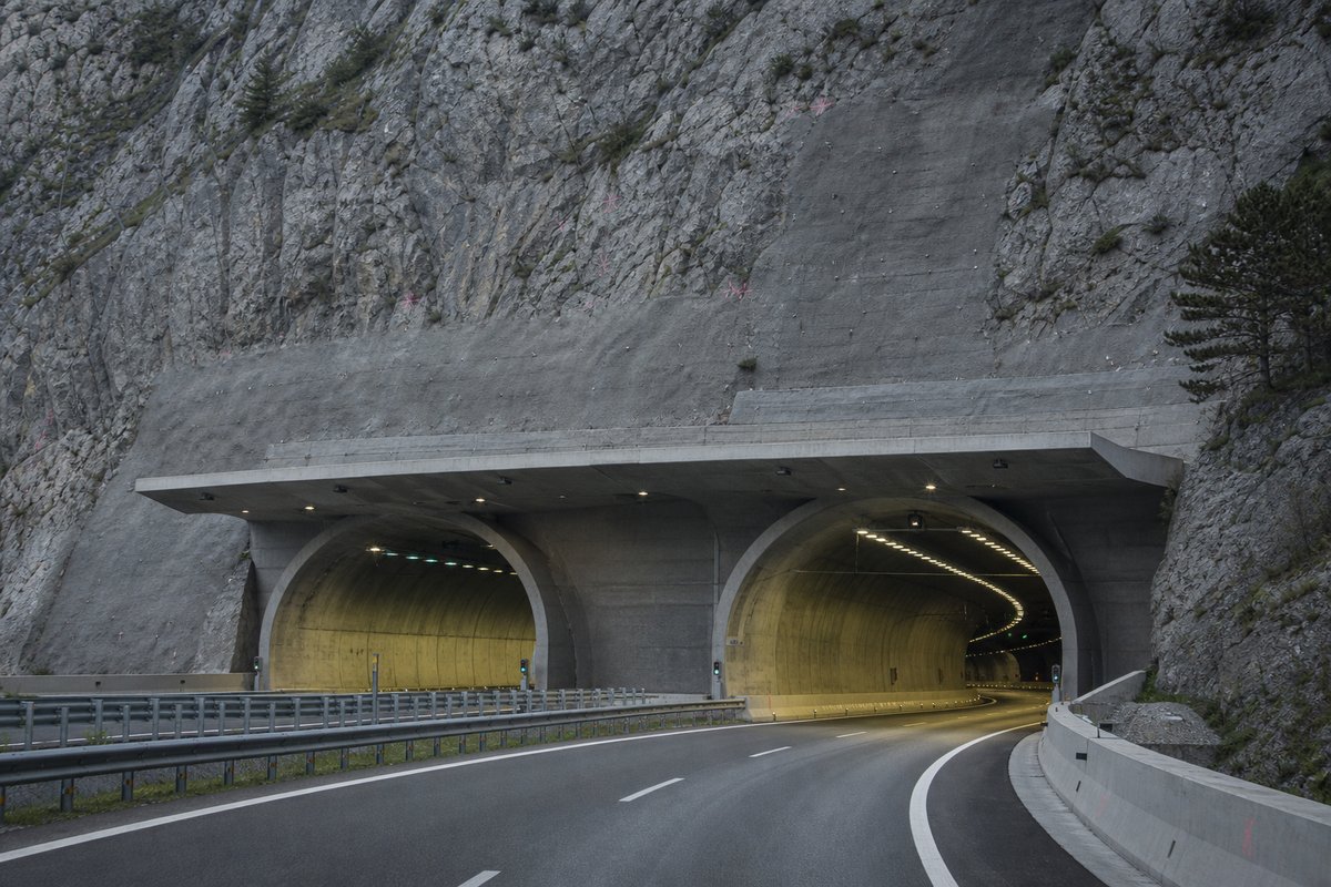 Road tunnel portal in Italy: Apennine limestone ridge, twin concrete motorway portals with a projecting canopy, in flat overcast light, shot at 85mm from a distance. (AI-generated image)