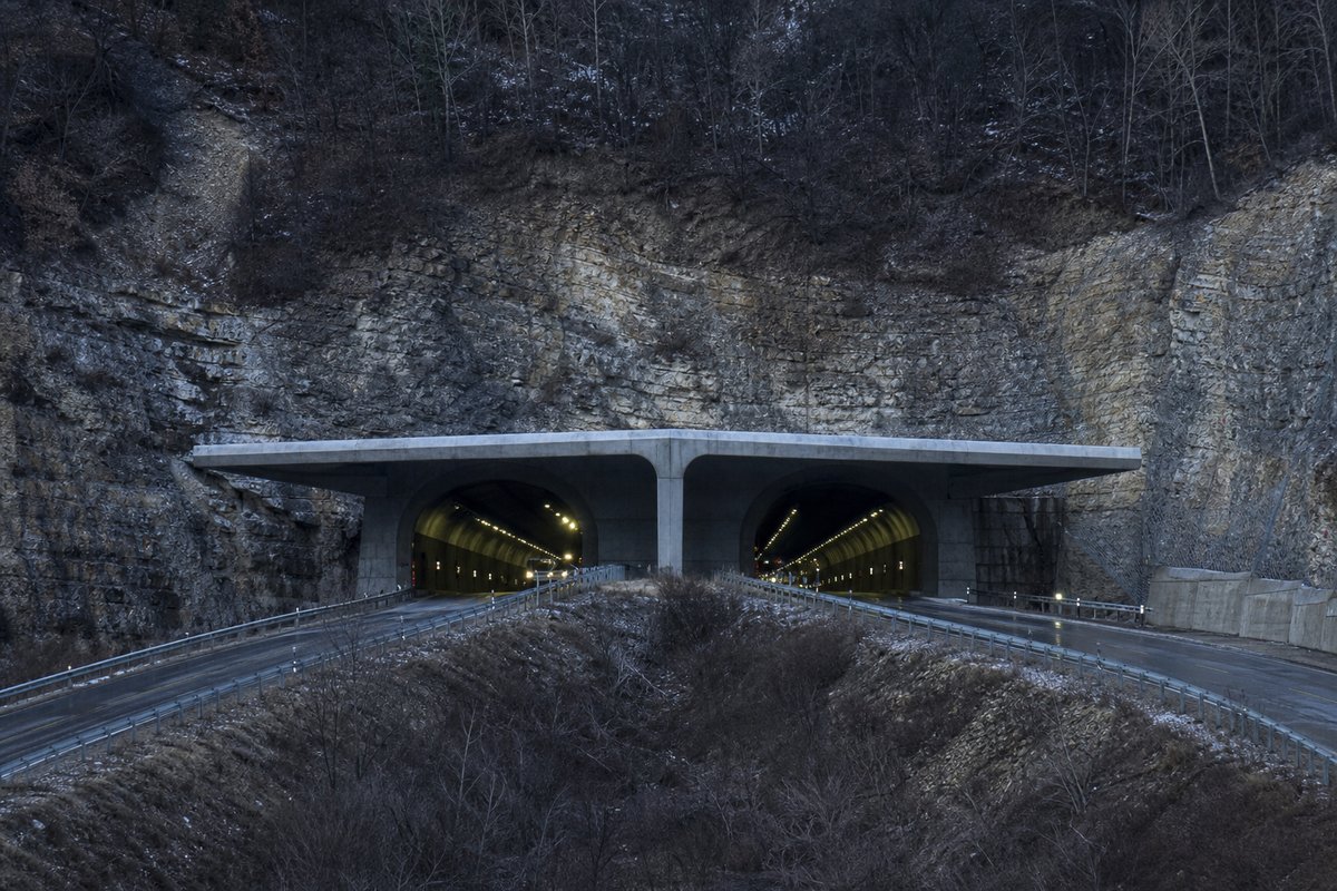 Road tunnel portal in Italy: Apennine limestone ridge, twin concrete motorway portals with a projecting canopy, in hard low winter sun, shot at 85mm from a distance. (AI-generated image)