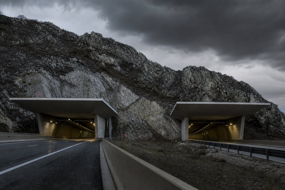 Transport infrastructure structure in Italy: Apennine limestone ridge, twin concrete motorway portals with a projecting canopy, under a heavy dark sky, shot at 50mm, compressed. (AI-generated image)