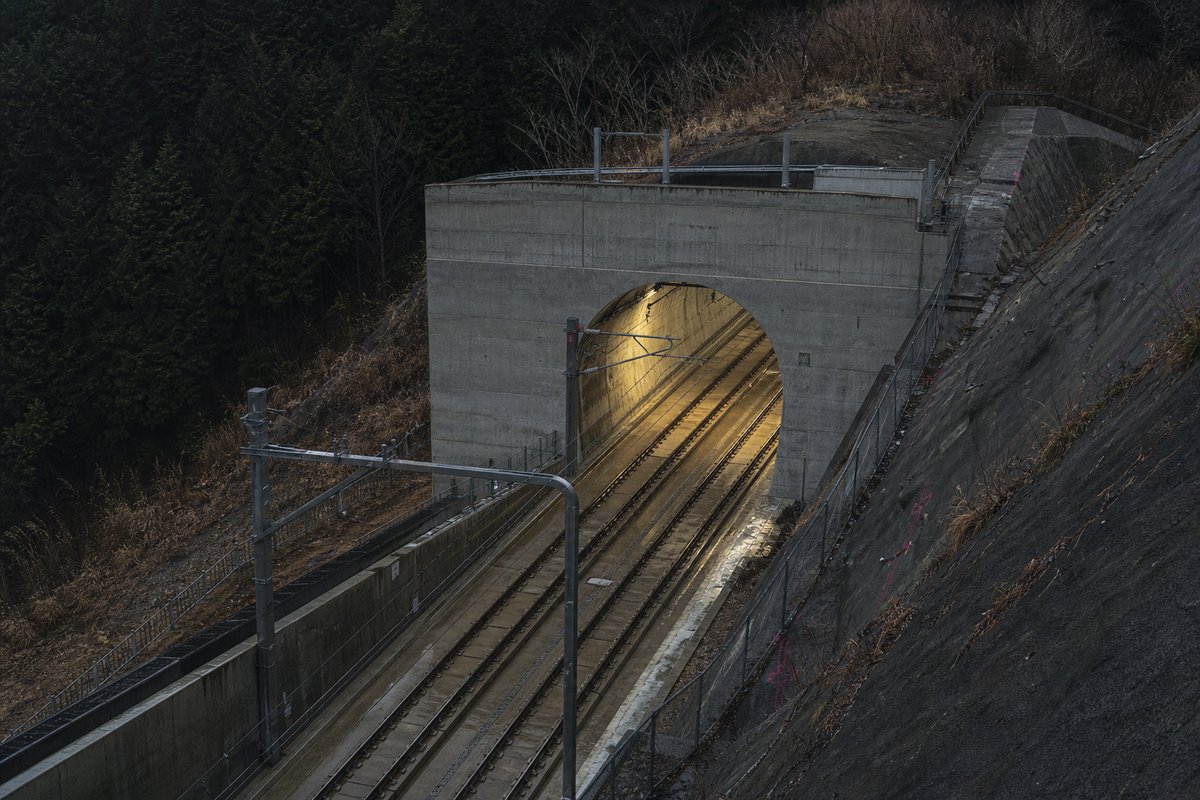 Transport infrastructure structure in Japan: forested volcanic ridge, a precise concrete rail portal with catenary wires, in hard low winter sun. (AI-generated image)