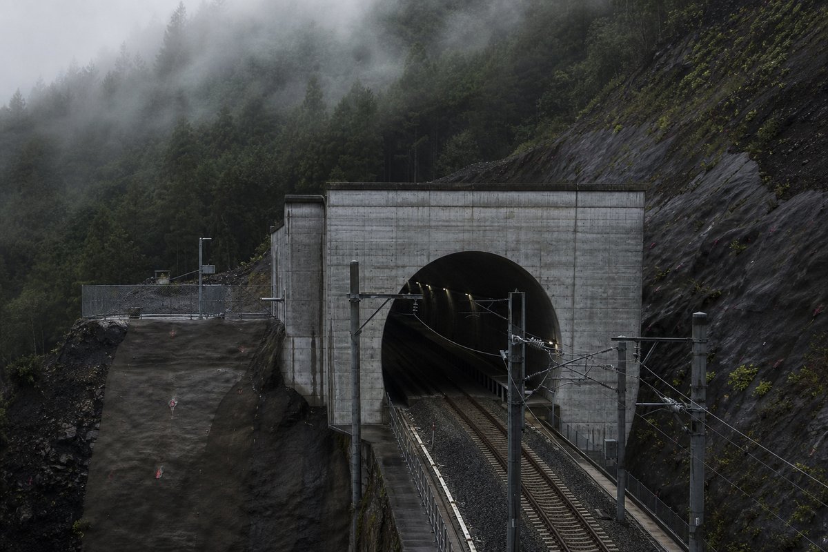 Road tunnel portal in Japan: forested volcanic ridge, a precise concrete rail portal with catenary wires, in thin morning fog, shot at 85mm from a distance. (AI-generated image)