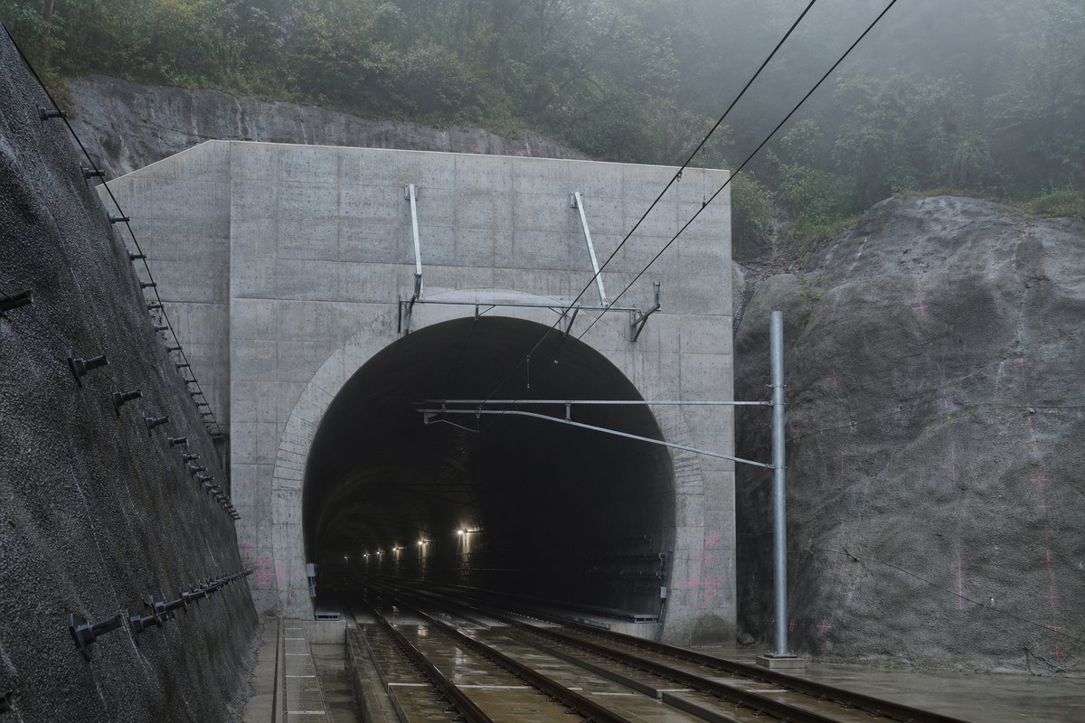Road tunnel portal in Japan: forested volcanic ridge, a precise concrete rail portal with catenary wires, in thin morning fog, shot at 35mm. (AI-generated image)