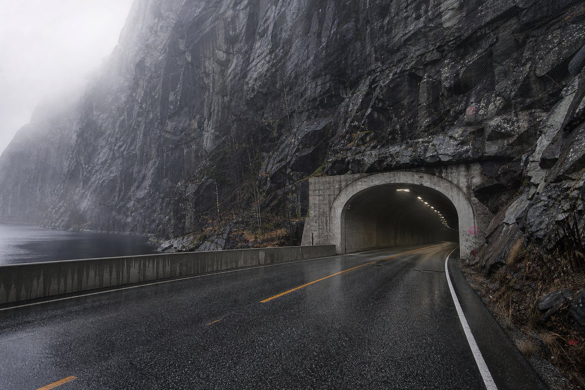 Transport infrastructure structure in Norway: steep granite fjord wall, dark water below, a rock-cut tunnel portal with no decoration, in thin morning fog. (AI-generated image)