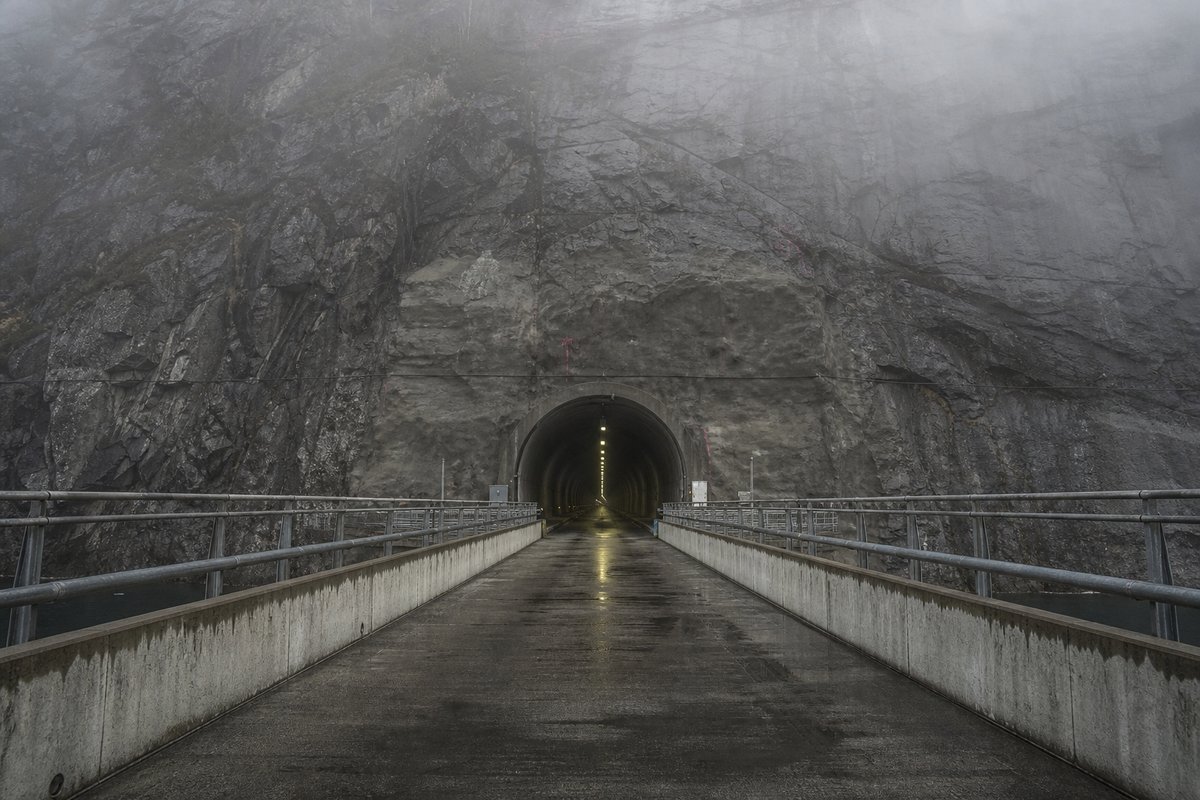 Road tunnel portal in Norway: steep granite fjord wall, dark water below, a rock-cut tunnel portal with no decoration, in thin morning fog, a dead-centre symmetrical composition. (AI-generated image)