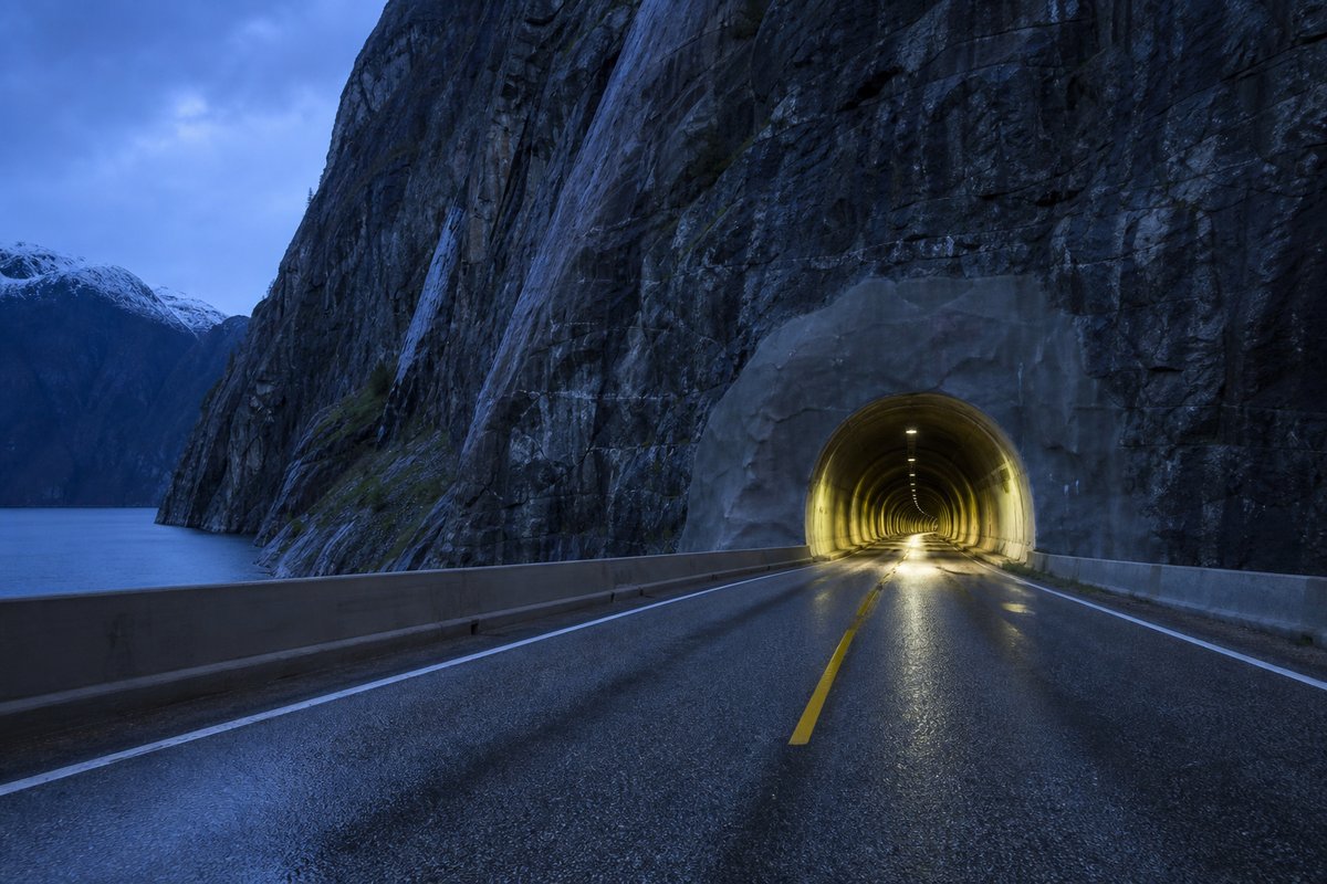 Road tunnel portal in Norway: steep granite fjord wall, dark water below, a rock-cut tunnel portal with no decoration, at blue hour with the portal lit. (AI-generated image)