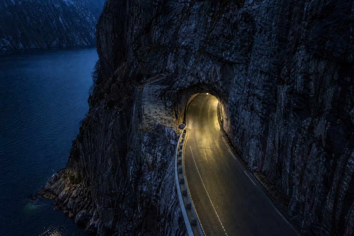 Road tunnel portal in Norway: steep granite fjord wall, dark water below, a rock-cut tunnel portal with no decoration, at blue hour with the portal lit. (AI-generated image)