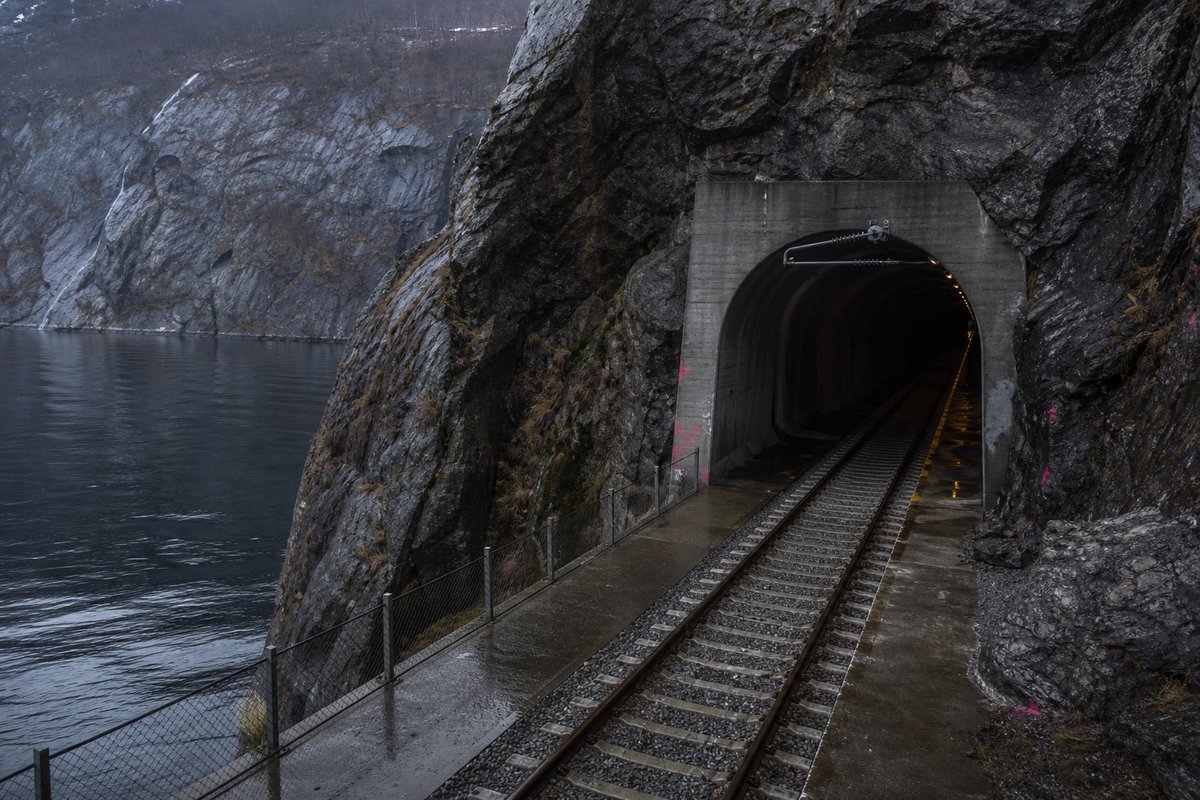 Railway tunnel portal with track running in in Norway: steep granite fjord wall, dark water below, a rock-cut tunnel portal with no decoration, after rain. (AI-generated image)
