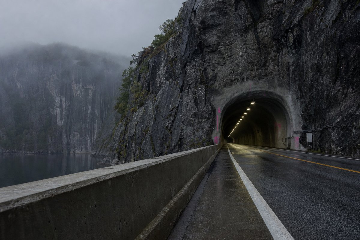 Transport infrastructure structure in Norway: steep granite fjord wall, dark water below, a rock-cut tunnel portal with no decoration, in thin morning fog. (AI-generated image)