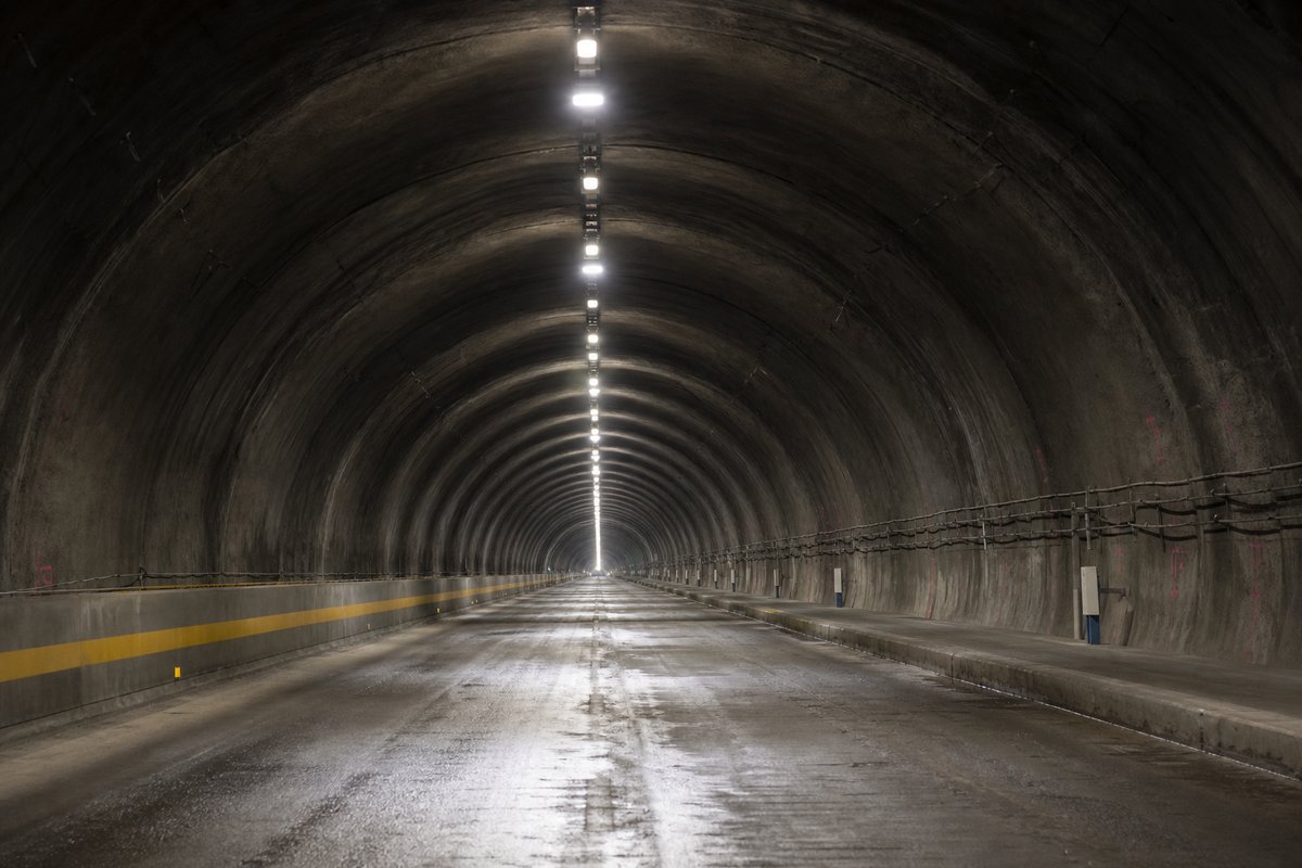 Looking down the axis of an extremely long two-lane road tunnel bore, luminaires receding to a point: a mountainous transport corridor, in flat overcast light, shot at 50mm. (AI-generated image)