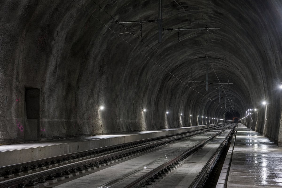 Double-track railway tunnel interior with ballastless slab track and catenary receding into darkness: a mountainous transport corridor, after rain, everything wet and reflective. (AI-generated image)