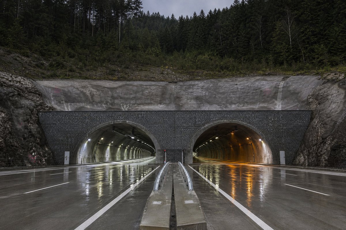 Road tunnel portal in Slovakia: Carpathian forested ridge with a new twin-tube portal, after rain, everything wet and reflective, a dead-centre symmetrical composition. (AI-generated image)