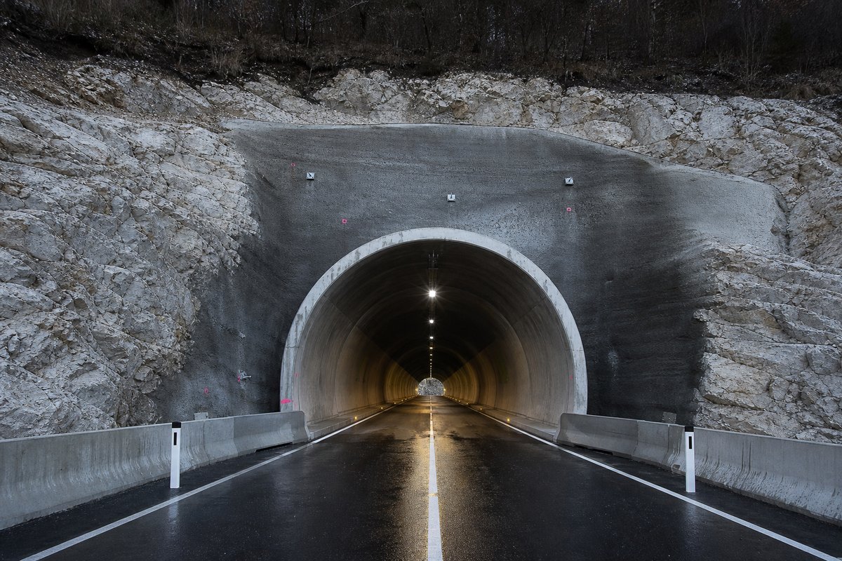 Road tunnel portal in Slovenia: karst plateau, a short tunnel through pale limestone, in hard low winter sun, a dead-centre symmetrical composition. (AI-generated image)