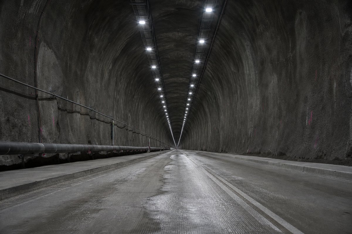 Looking down the axis of an extremely long two-lane road tunnel bore, luminaires receding to a point: a mountainous transport corridor, in flat overcast light. (AI-generated image)